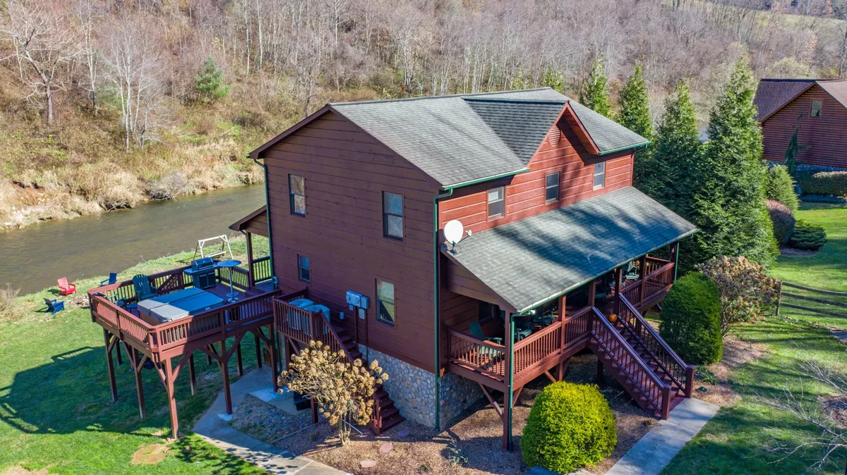 This exterior image features a two-story wooden house with a spacious deck that includes a hot tub. The river can be seen in the background, enhancing the sense of direct water access. The setting highlights outdoor relaxation and is framed by natural surroundings, perfect for both quiet moments and group gatherings.