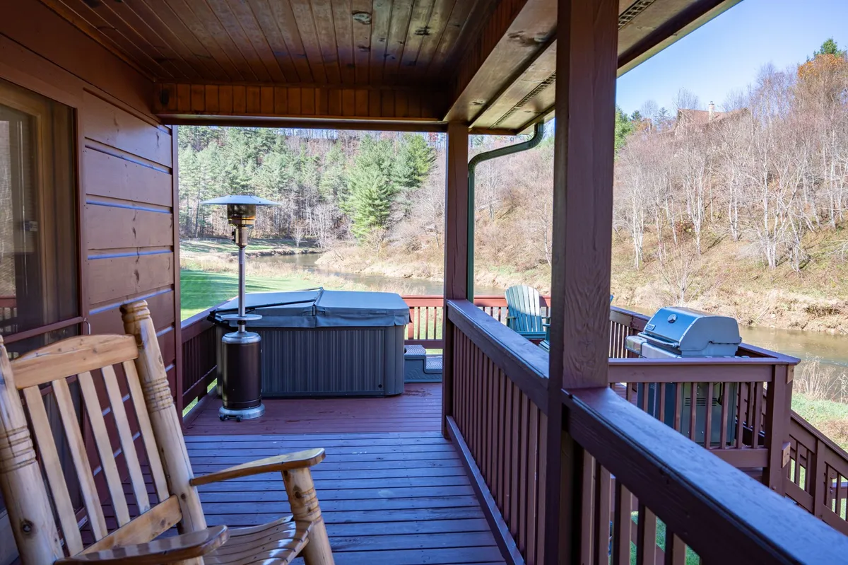 A broad deck with a rocking chair, hot tub, outdoor grill, and a panoramic view of the river framed by green trees. The setup features spaces for relaxation, dining, and soaking, all while enjoying the sights and sounds of the surrounding mountains and waterway.