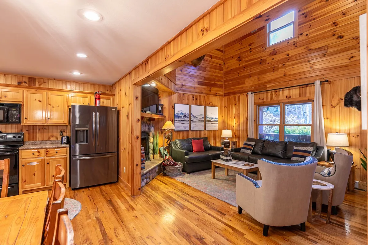A view of the open living room and kitchen area, featuring inviting sofas, a functional kitchen space with modern appliances, a fireplace, and artwork on the walls. The open layout encourages interaction between cooking, dining, and relaxing.