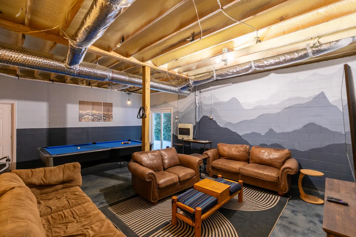 A spacious game room featuring a pool table at the center, surrounded by luxurious leather sofas and a striking mountain mural on the wall. The relaxed atmosphere is ideal for both casual gatherings and more competitive game nights. The room is designed for both recreation and socializing, with ample seating for groups.