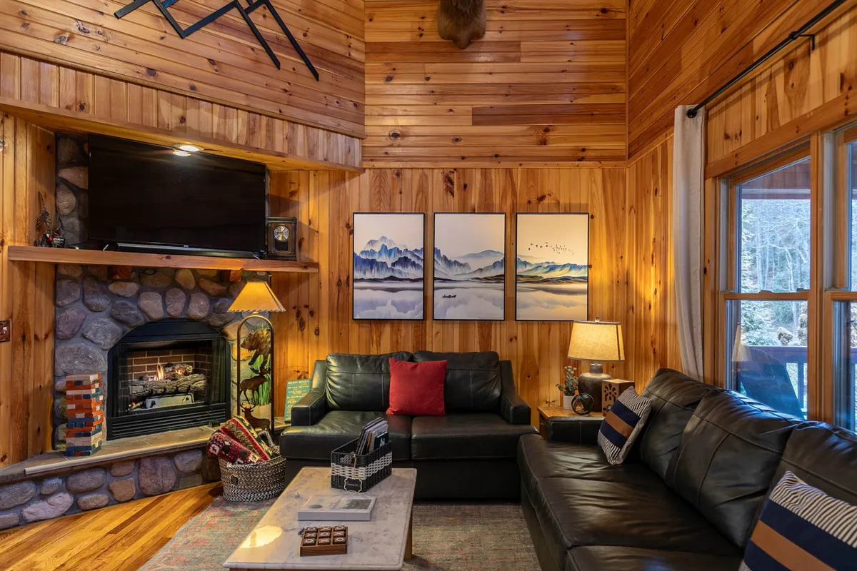 A warm and inviting interior scene featuring a cozy gas-log fireplace, comfortable sofas, soft lamp lighting, and tasteful wall art. The setup suggests a space ideal for relaxing by the fire, reading, or sharing laughs during game or movie nights.