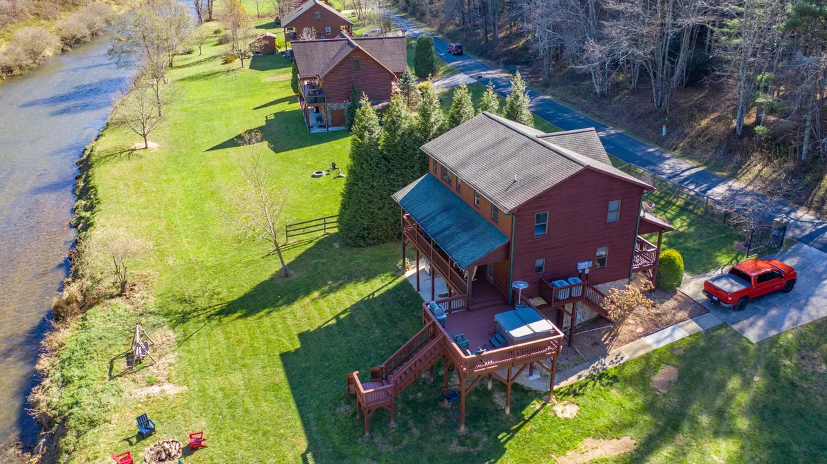 A wide view of the river, green lawn, fire pit, and deck, with houses in the background. The expansive yard leads directly to the water, demonstrating easy, open access for recreation and play. The fire pit and deck are visible, supporting social activities.