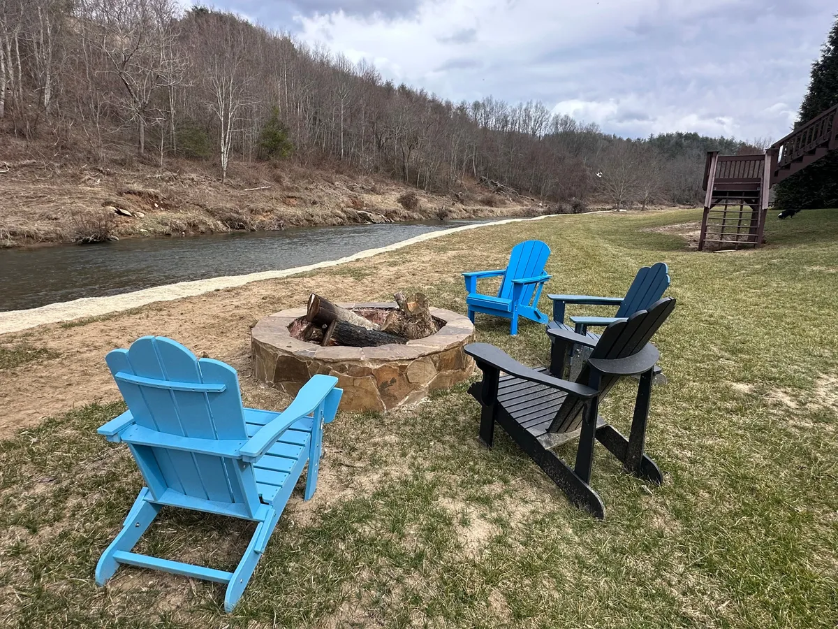 A tranquil fire pit area with Adirondack chairs faces the river, surrounded by greenery. The fire pit is the centerpiece, inviting guests to gather for s'mores or storytelling as night falls, while the river and surrounding landscape complete the inviting outdoor scene.