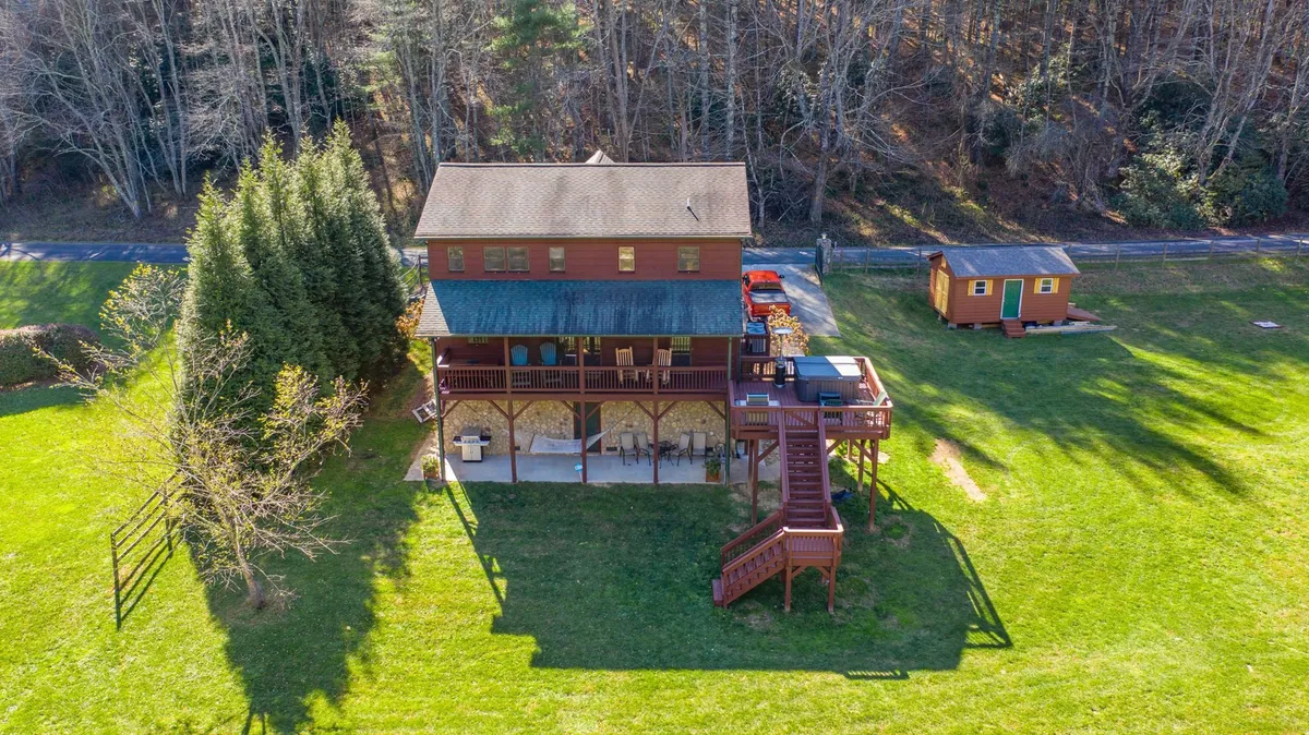 A wide-angle view of the two-story wooden house, showcasing a spacious deck, an expansive green lawn, and surrounding trees. The image hints at the outdoor possibilities with visible play areas for children and ample space for gatherings or lawn games.