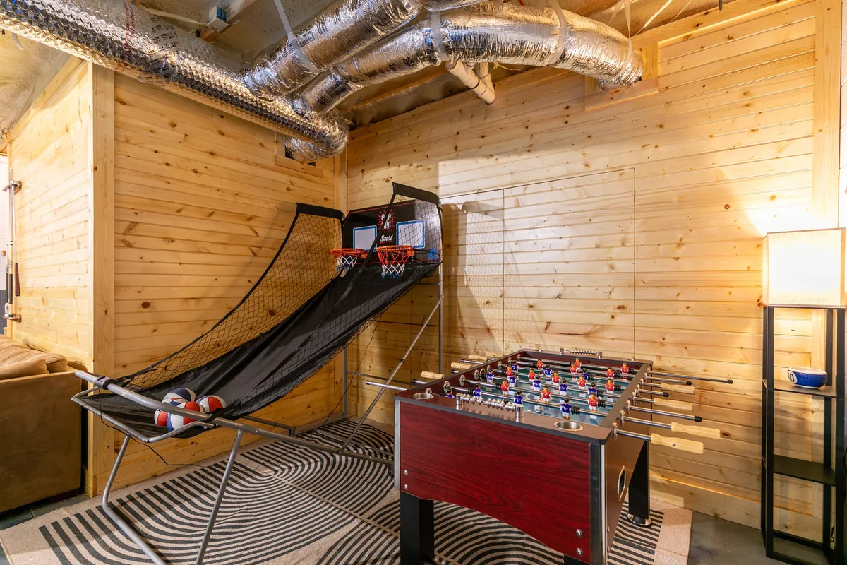A lively corner of the game room with a foosball table, basketball arcade game, and striped rug set against wooden walls. The space feels fun and competitive, inviting guests to engage in multiple games at once.