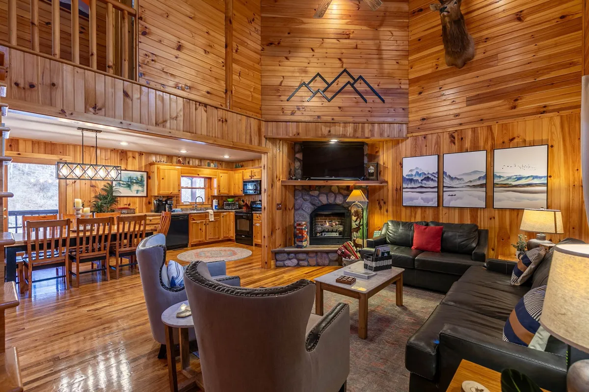 This image presents a spacious, open-concept living area with a central fireplace as the focal point. Large windows let in natural light, while a wooden dining table and mountain-themed artwork add a warm, rustic touch. The seating is generous, inviting gatherings for family and friends. The visible kitchen elements in the background emphasize the open, connected nature of the primary living space.